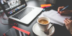 cup of coffee on a plate with spoon with open laptop and notebook in backgroupnd